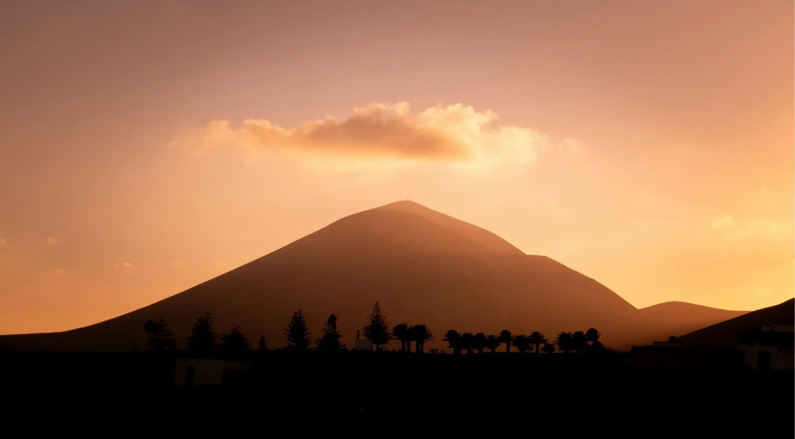 A volcanic cone in silhouette at sunset above a line of palm trees
