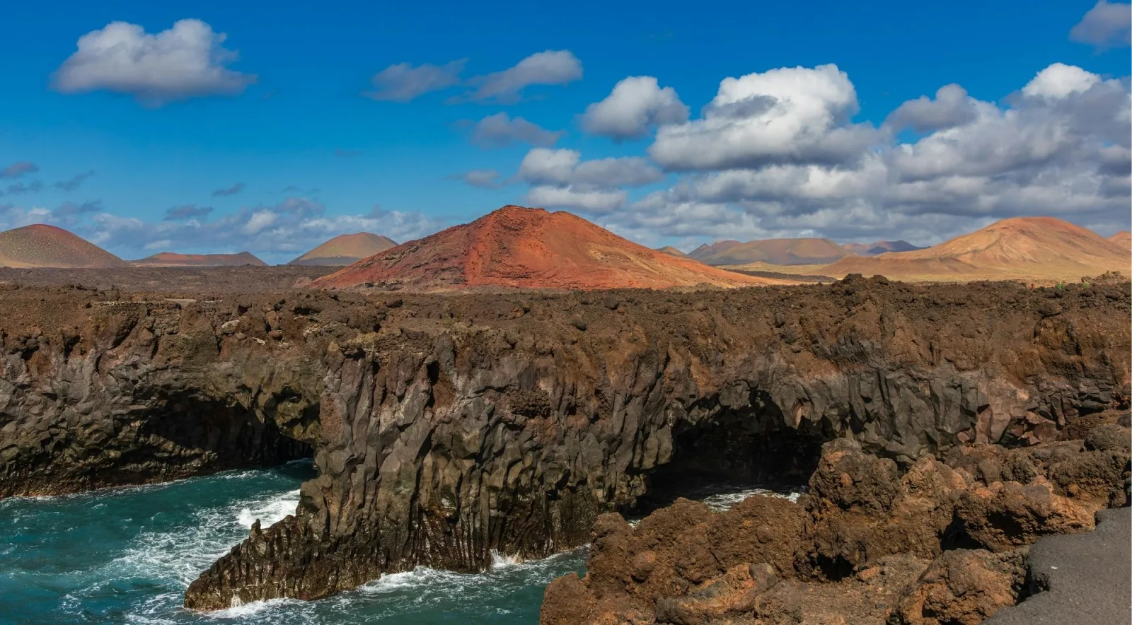 Waves breaking against the black lava cliffs of Los Hervideros with a red volcanic cone inland