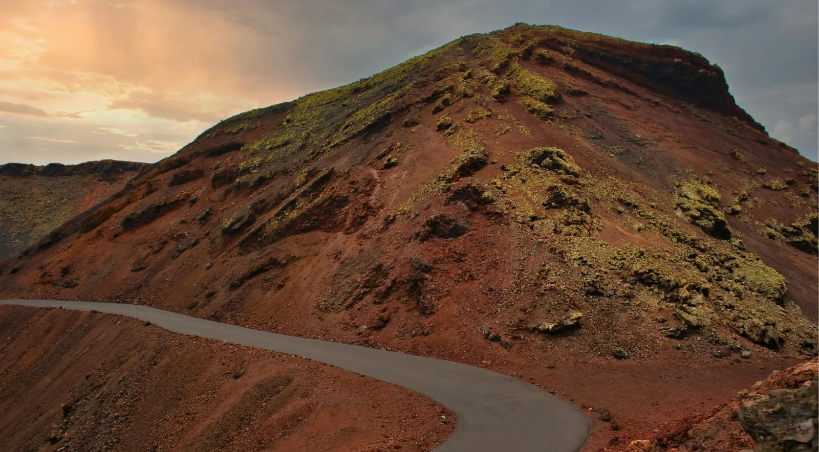 A narrow road curving around a red-brown volcanic slope at dusk, lichen patching the cinder
