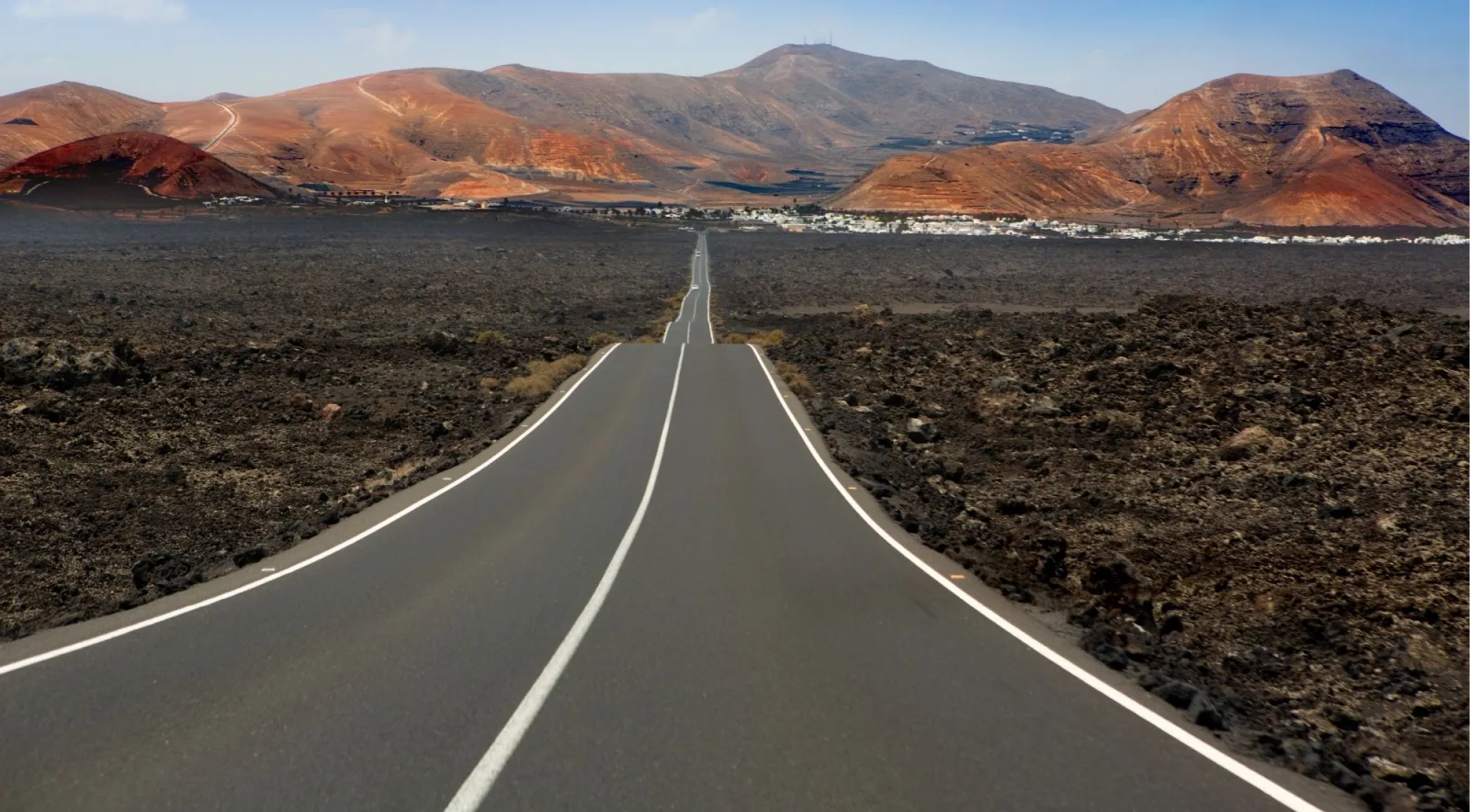 A straight road crossing a black lava field towards Lanzarote's red-brown hills