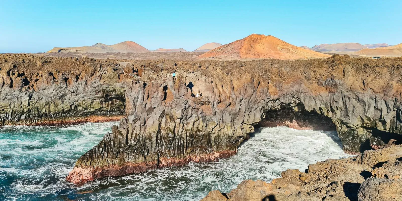 Waves breaking through the eroded lava arches of Los Hervideros below a red volcanic cone
