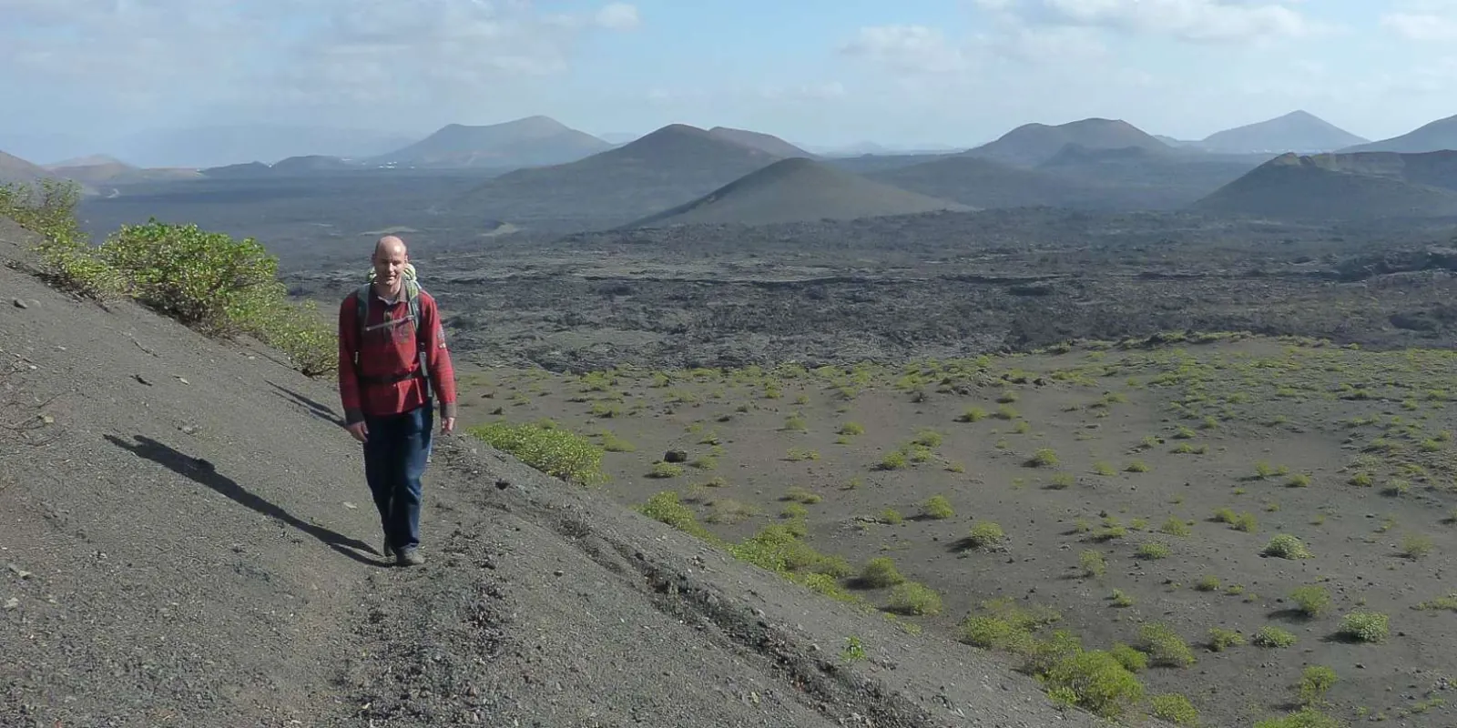 A hiker in a red jacket on a cinder ridge above the hardened lava sea, cones on the horizon
