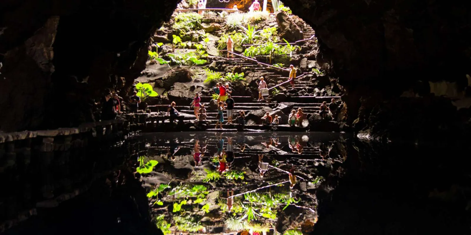 Visitors on the lit stone terraces inside the Jameos del Agua cave, reflected in the pool